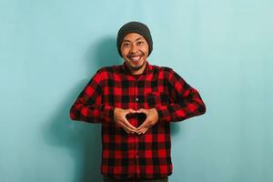 Smiling young Asian man with a beanie hat and a red plaid flannel shirt shows a love gesture with his hand while standing against a blue background photo