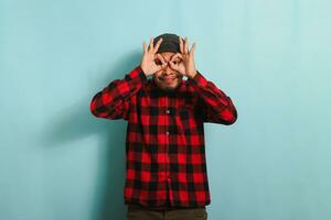 Excited young Asian man with beanie hat and red plaid flannel shirt makes the OK sign gesture with his fingers, acting as binoculars with his eyes peeking through them, isolated on blue background photo
