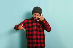 An excited young Asian man wearing a beanie hat and a red plaid flannel shirt makes a call me gesture, pointing down to the copyspace, isolated on a blue background photo