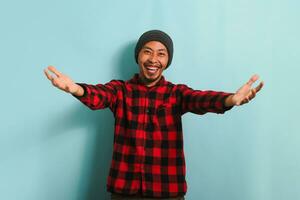 sonriente joven asiático hombre vistiendo un gorro sombrero y un rojo tartán franela camisa es llamando con ambos manos, ofrecimiento un abrazo con un ven aquí gesto, atractivo, acogedor, aislado en azul antecedentes foto