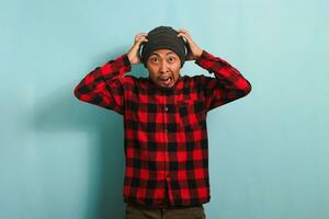 Scared Young Asian man with a beanie hat and red plaid flannel shirt has an open mouth, grabs his head, and looks at the camera reacting to something horrible, isolated on a blue background photo