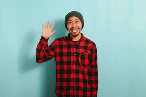 An attractive and friendly young Asian man with a beanie hat and a red plaid flannel shirt smiles, waves, and says hello, hi, or bye while standing against a blue background photo