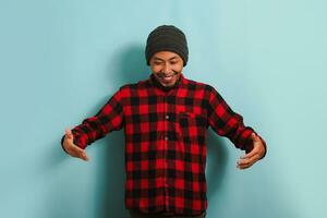 Young Asian man with beanie hat and red plaid flannel shirt gesturing with hands and showing a big and large size sign, representing a measurement symbol, isolated on a blue background photo