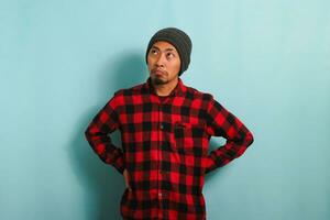 A bored young Asian man wearing a beanie hat and a red plaid flannel shirt stands with an arm akimbo gesture, looking to the left deep in thought about what to do, isolated on a blue background photo