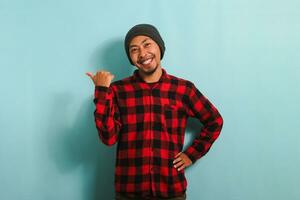 emocionado joven asiático hombre con un gorro sombrero y rojo tartán franela camisa es señalando aparte con su pulgar, indicando el Copiar espacio, aislado en un azul antecedentes foto