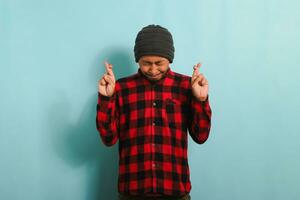 A hopeful young Asian man with a beanie hat and a red plaid flannel shirt makes a finger crossed gesture, closes his eyes, and makes a wish while standing against a blue background photo