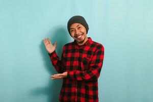 An excited Young Asian man with a beanie hat and a red plaid flannel shirt is showing with his hands to the side, indicating the empty copy space, isolated on a blue background photo