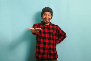un sonriente joven asiático hombre con un gorro sombrero y un rojo tartán franela camisa extiende su mano para un apretón de manos, acogedor y ofrecimiento cooperación mientras en pie en contra un azul antecedentes foto