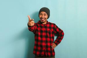 Excited Young Asian man with beanie hat and red plaid flannel shirt is smiling and pointing his finger at the camera, choosing or picking, and inviting YOU to join, isolated on blue background photo