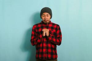 A young Asian man with a beanie hat and a red plaid flannel shirt folds his hands in prayer to God while standing against a blue background photo