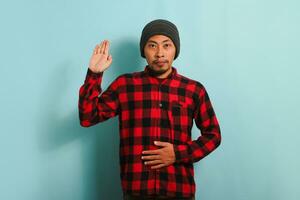 A young Asian man with a beanie hat and red plaid flannel shirt is swearing with his hand on his chest and an open palm, making a loyalty promise oath, isolated on a blue background photo