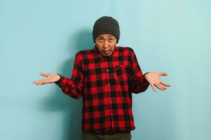 A confused young Asian man with a beanie hat and a red plaid flannel shirt is shrugging his shoulders and looking at the camera with a puzzled expression, isolated on a blue background. photo