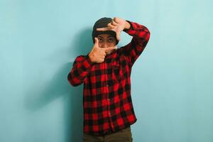 emocionado joven asiático hombre con un gorro sombrero y un rojo tartán franela camisa es haciendo un imagen marco gesto, fingiendo a atención como un fotógrafo, aislado en un azul antecedentes foto