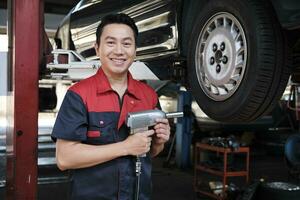 One expert Asian male automotive mechanic technician is screwing car wheel nuts on lifting with drill wrench for repair at garage. Vehicle maintenance service works industry occupation business jobs. photo