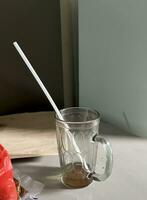 Finished empty sweet iced tea liquid drink beverage in clear glass mug cup with white straw inside. Isolated on table top with gray walls and visible sun light, and red plastic object beside it. photo