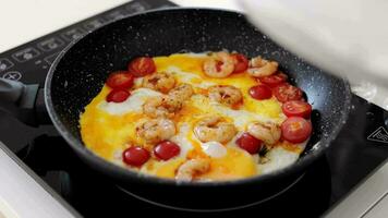 omelete com cereja tomates, legumes e camarão dentro uma panela às lar. mulher cozinhando mexidos ovos em elétrico fogão topo dentro a cozinha, fritar panela com frito ovos em indução cooktop fechar-se. video