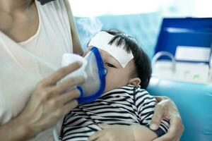 madre Ayudar pequeño hijo utilizando nebulizador durante inhalación. foto