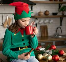 Child in elf costume holding chocolate biscuit and mug with candy cane in it in hands photo