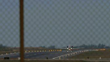 A private jet with an unrecognizable livery speeds up for takeoff at dusk. View through the airport fence. Landing lights, lighting, runway video