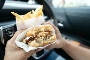 asiático dama participación hamburguesa y francés papas fritas a comer en auto, peligroso y riesgo un accidente. foto