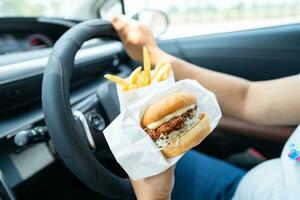 Asian lady holding hamburger and French fries to eat in car, dangerous and risk an accident. photo