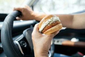 Asian lady holding hamburger to eat in car, dangerous and risk an accident. photo