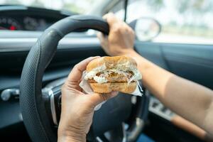 asiático dama participación hamburguesa a comer en auto, peligroso y riesgo un accidente. foto