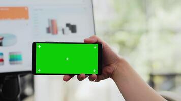 Specialist looks at greenscreen display on mobile phone, holding gadget with copyspace isolated software at workstation. Employee working with blank mockup layout in business office. Close up. video