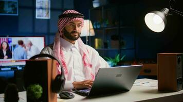 glücklich Arabisch Telearbeiter antworten Job E-Mails im stilvoll Wohnung. heiter Muslim Mitarbeiter entfernt Arbeiten, Tippen Daten auf Laptop mit geöffnet Fernseher wie Hintergrund Lärm, Handheld Kamera Schuss video