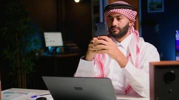 retrato de contento Arábica teletrabajador enviando texto mensajes a amigos mientras trabajando desde hogar en elegante departamento. alegre medio oriental hombre mecanografía en móvil teléfono con abrió televisión como antecedentes ruido video