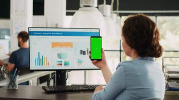 Employee looks at greenscreen on phone, working on business operations at desk. Person holding smartphone device with isolated display showing mockup template, blank layout screen. video