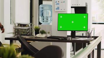 Empty workstation with greenscreen running on computer, open floor plan coworking space presenting isolated display with chroamkey template. Monitor on desk showing blank copyspace layout. video