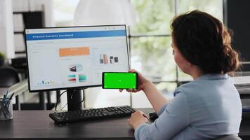 Businesswoman looks at greenscreen while she holds mobile device, isolated chromakey display shown at office desk. Employee checking blank copyspace software on phone. Handheld shot. video