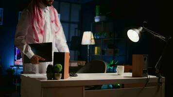 Happy Arabic employee arriving in stylish apartment personal office, starting important job tasks. Smiling Middle Eastern teleworker typing on laptop keyboard, beginning work shift at home video