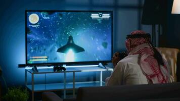 Arabic gamer playing classic arcade action videogame, flying through space debris. Muslim man enjoying leisure time at home using high tech gaming console to solve missions in singleplayer game video