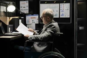 Professional accountant in wheelchair checking administrative paperwork in dark file room. Businessman in bureaucratic file cabinet office filled with invoice folders and flowcharts photo