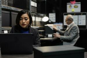 Professional business workers checking administrative analytical data in bureaucratic office filled with invoice folders. Dark file room surrounded by accountancy statistics flowcharts. photo