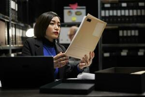 Asian private detective in agency file cabinet room reading criminal case file details. Investigator woman surrounded by criminology document folders on office repository cabinet shelves photo