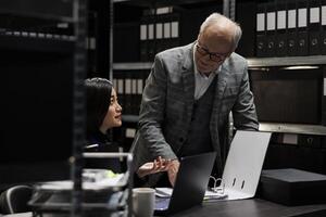 Business invoicing administration employees discussing bookkeeping data analysis paperwork. Archival storage room office filled with flowcharts and document folders on cabinet shelves photo