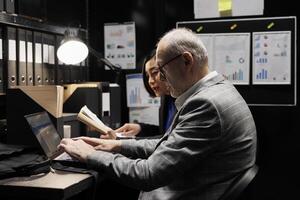 Professional elderly businessman manager and asian assistant checking administrative analytical data in file cabinet office. Accounting team in bureaucratic office filled with invoice folders photo