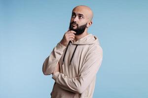 Young arab man touching chin thoughtfully and looking away with doubt facial expression. Person wearing casual clothes while thinking and solving problem with pensive emotions photo