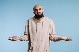 Arab man with spread arms and puzzled expression showing do not know gesture portrait. Young bald beared person looking at camera with confused emotions while shrugging shoulders photo