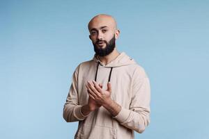 Arab man applauding with hands while standing and looking at camera with neutral expression. Young bald bearded person clapping with arms, congratulating and supporting studio portrait photo