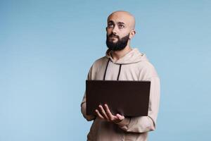 Thoughtful arab man holding laptop and looking upwards with pensive expression. Young person in doubt programming software application while standing with portable computer photo