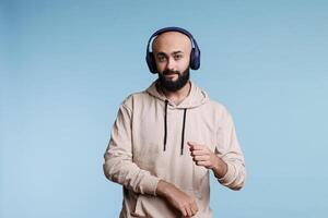 Arab man listening to music in headphones and making moves with hands portrait. Relaxed young person wearing wireless earphones dancing while enjoying song and looking at camera photo