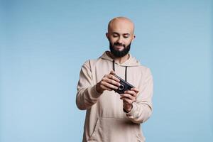 Young arab man playing mobile game, using entertaining application software. Concentrated gamer enjoying videogame on smartphone while standing in studio on blue background photo