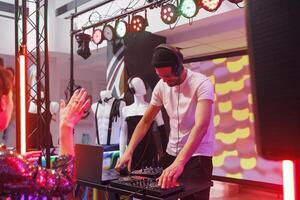 Smiling musician mixing electronic tracks with dj station for people partying in nightclub. Young man performing on stage while clubbers having fun and dancing in club with spotlights photo