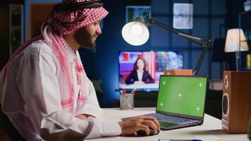 Muslim man scrolling on isolated screen laptop, checking social media posts. Self employed person browsing internet websites from chroma key device in apartment office with TV as background noise video