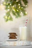 Milk for Santa. Glass milk, and cookies on ,wooden stand. Fir branch, Christmas lights on white wall photo