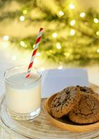 galletas y Leche en vaso en blanco redondo de madera pararse. abeto rama, Navidad luces. Leche para Papa Noel foto